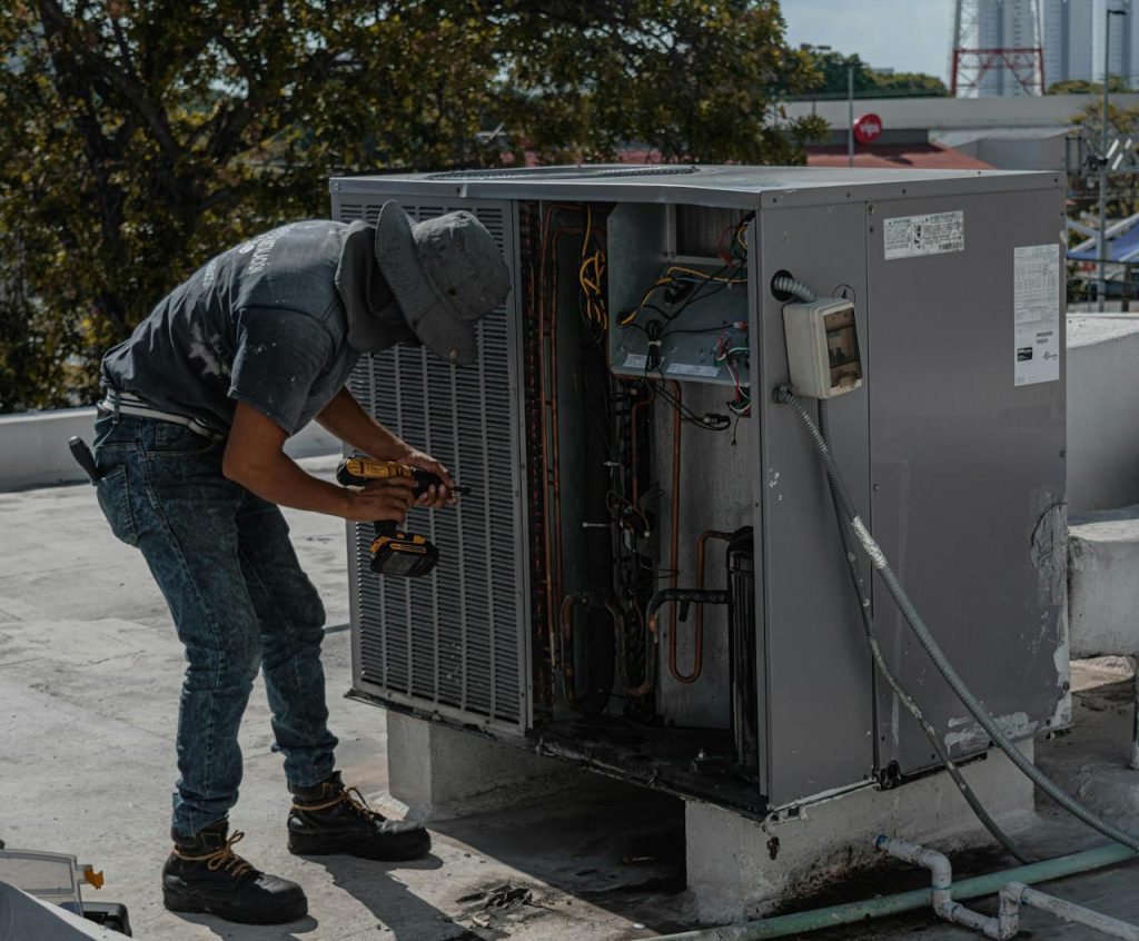 NR Electric Job 3 A worker in a bucket hat repairs an outdoor air conditioning unit on a rooftop.